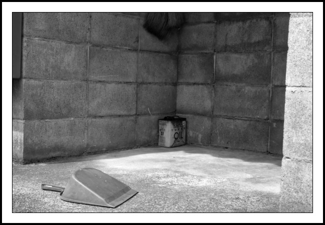 A black and white photograph of a dustpan, an old tin of oil and the bristles of a broom. These three objects can be found inside a three-sided concrete block structure: The dustpan in the opening of the three sided structure, the broom bristles (top middle of the frame) are in the left corner of the structure hanging ominously above the oil tin which fits tightly into that same corner on the floor. There is light coming in from the left hand side of the photograph but the oil tin and the broom bristles are still somewhat shaded. The dustpan is in full light - bottom left of the photograph. The dustpan is upturned? Why? And where is its brush? The bristles of the broom are large and would suggest a long handle based means of use. Sure you could do the initial sweeping with those bristles but it will take a little work to then use the same broom to deposit the swept dust into the dustpan. As for the tin of oil it’s real specific: Daihatsu Mira Turbo Oil. The Mira was in production from 1980 until 2018, so that is an old tin cowering in the corner. The whole scene is very tidy (and free of dust) so I guess that some of the items have been used recently. However, the oil tin looks trapped, unused and forgotten. Have the dustpan and broom bristles been ganging up on it? Do we have a Mexican standoff in progress?