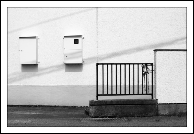 A black and white 3:2 ratio landscape orientated photograph of a white textured concrete building wall with two boxes affixed thereto. A black metal fence with two ropes tied and a white concrete wall are also included in the photograph. The two boxes are in the left third of the frame with their bases about halfway up and their tops about a quarter of the way down. I guess the boxes are electrical in function. They both have small Yale-style locks midway on their left-hand side. The left hand box - which is close to the left edge of the photo - is completely encased in metal. The right-side one - the right side of which is relatively close to the centre of the image - has a small darkened window in its top right. Otherwise it too is clad in metal. Both boxes exude a semi-circle glow above them which will be fun to play with when I do a full edit on the photograph. The black fence has 12 rungs inside its frame. The left of the frame is exactly halfway along the photo and lines up with a depression in the back wall. This was the main reason return and so they lined-up better. The top of the fence frame is halfway up from the bottom (or halfway down from the top if you prefer). Two ropes have been tied to the fence: the top rope to the eleventh rung and the bottom rope to the twelfth rung. The ropes are both taut and run to the right of the photograph but are hidden by the white concrete wall which starts very close to the right frame of the fence. That wall runs out of the photo (not literally) on the right and is a little higher than the height of the fence. The fence sits on a sturdy concrete base as does the wall. These bases are separate constructions but joined together: unlike what they are supporting. The fence base is a more darker colour than the wall’s - perhaps it doesn’t take the weathering that well? There are two overhead cable shadows cast along the wall. The lower one runs the complete width of the photograph. It starts bottom left just a tad above the exposed ‘foundation layer’ of the building wall. This foundation’s top is about a third of the way up from the bottom of the frame. The shadow then slopes up and its bottom edge clips the top left corner of the fence frame and then clips the top left corner of the wall next to the fence before departing photograph stage right. The second, slightly weaker shadow, starts also on the left about a quarter of the way from the top edge of the photograph. Sloping upwards it clips the top of that central building wall depression exactly in the vertical centre of the photograph and at the top edge of the photograph. Yes, I needed to crop but I’m working with a 95% viewfinder. Allow me some wiggle room (about 5% methinks)? So after all that, what are the ropes doing then Guy? Well, I’m so glad you asked. Each prior visit I didn’t bother to look. So knowing that my eager audience would need to know I had a peep behind the curtain as it were… suffice to say the ropes are not holding up the fence but are helping to secure a couple of large objects to the right of the wall and well out of the frame of this photograph. Fret not, dear reader, a photograph of what those ropes support has been captured and will appear on the site within the month. In the meantime, feel free to let me know any suggestions you might have as to what those objects are. Until then…take care!