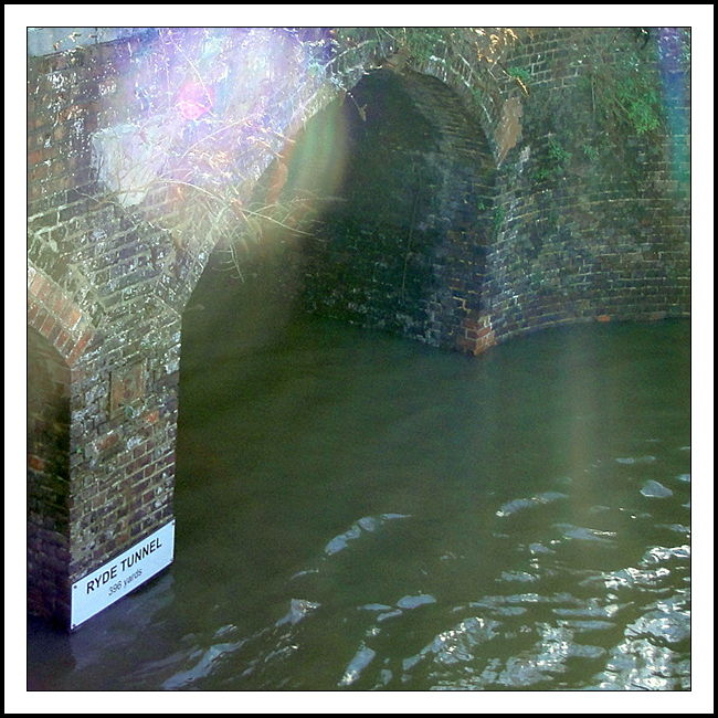 A photograph of the entrance to a tunnel through which water is flowing. The tunnel is named, as per a name board on the bottom left of the picture, the Ryde Tunnel