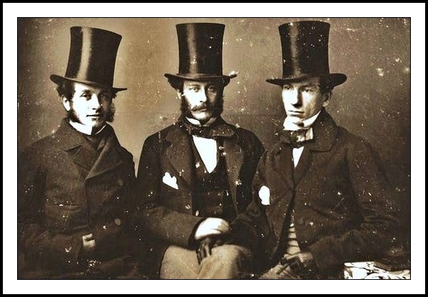 A vintage photograph of three typically styled Victorian men dressed up to the nines with matching, very tall, top hats: Stove-pipe top hats. The three men are sitting close together with the two men on the left side looking directly into the lens and at the viewer. The gentleman on the right is looking bottom left of the shot with a distant look in the eye. Methinks the gentleman would rather be somewhere else than stuck in a photographer’s studio